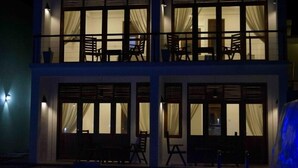 Un edificio de dos pisos con ventanas grandes, algunas con cortinas, y un balcón con sillas.