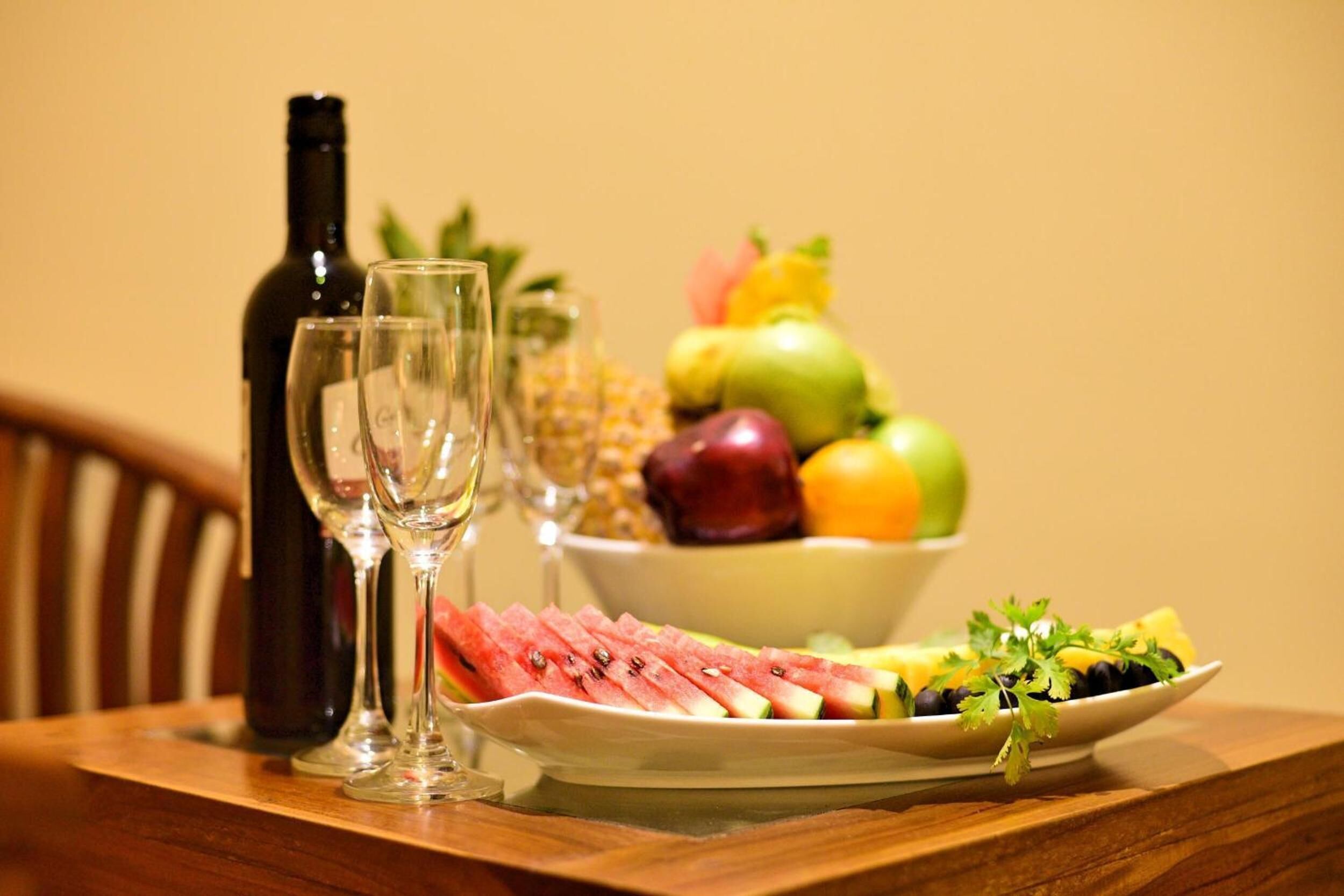 A dining table with a bottle of wine, two glasses, a bowl of fruit, and a plate of sliced watermelon and other fruits.