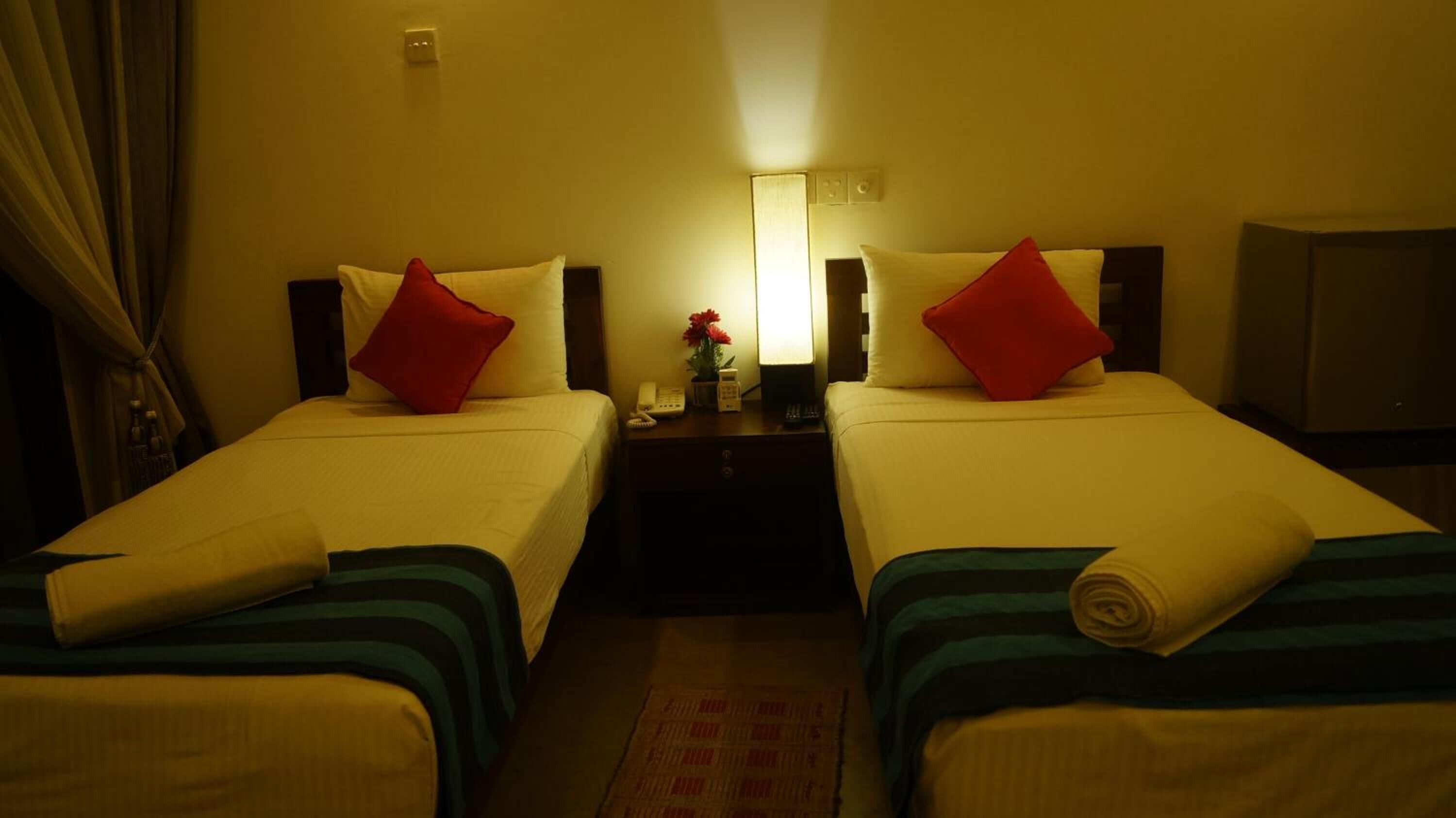 Quarto de hotel com duas camas de solteiro, cada uma com um travesseiro vermelho e um cobertor branco. Há uma mesa de cabeceira com uma luminária, um telefone e um vaso com flores.