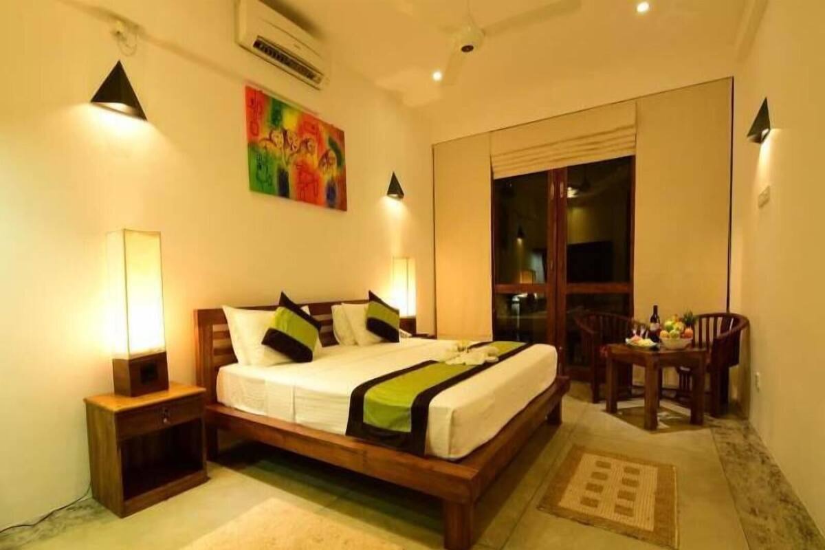 A hotel room with a bed, bedside tables, a window, a dining area, and a painting on the wall.