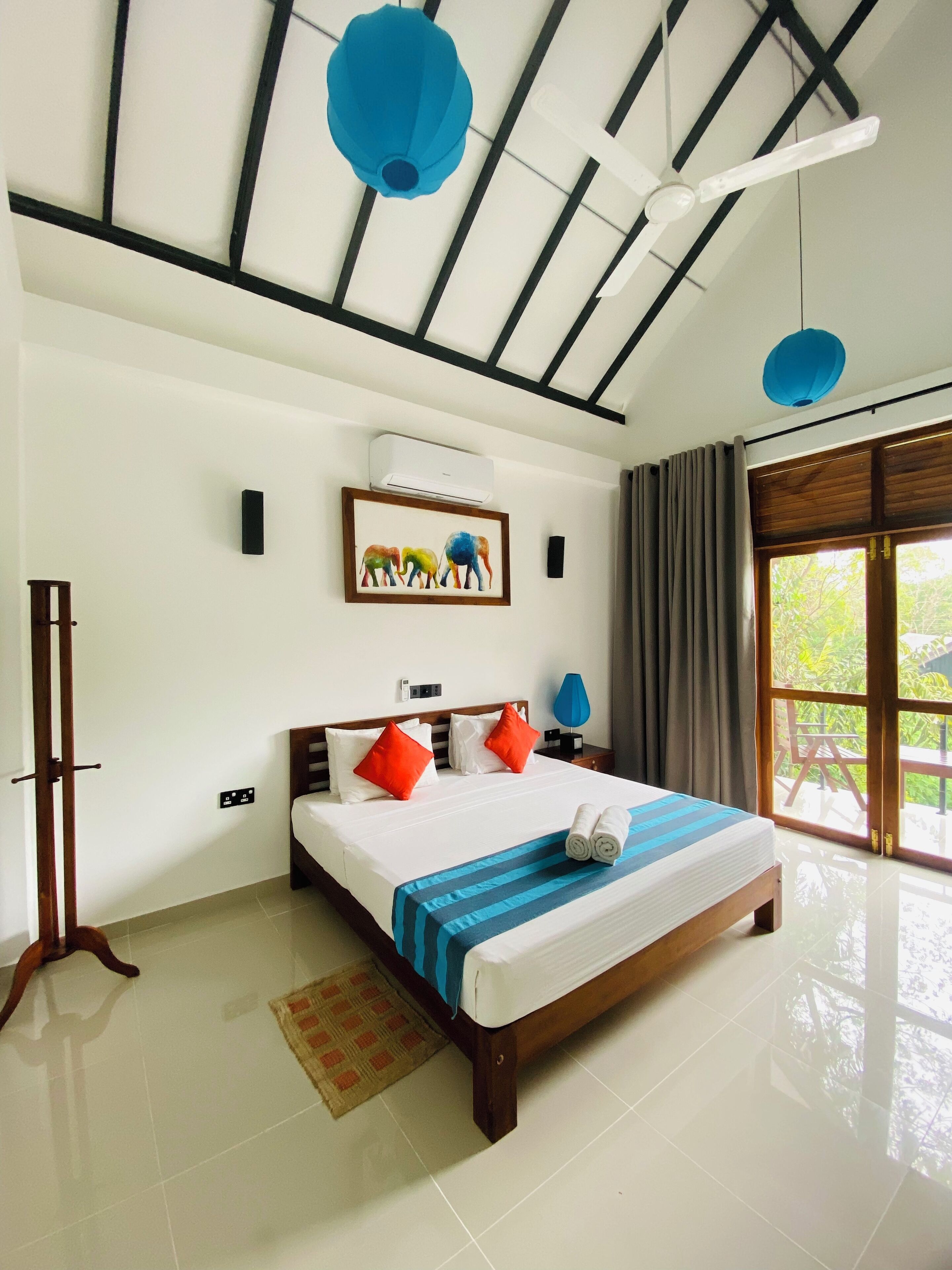 Habitación de hotel con una cama grande, un ventilador de techo, una ventana con cortinas y un cuadro de elefantes en la pared.