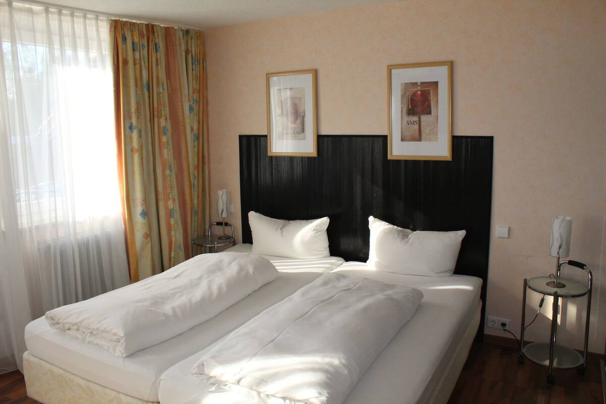 A hotel room with two beds, a headboard, a bedside table, a lamp, and a window with curtains.
