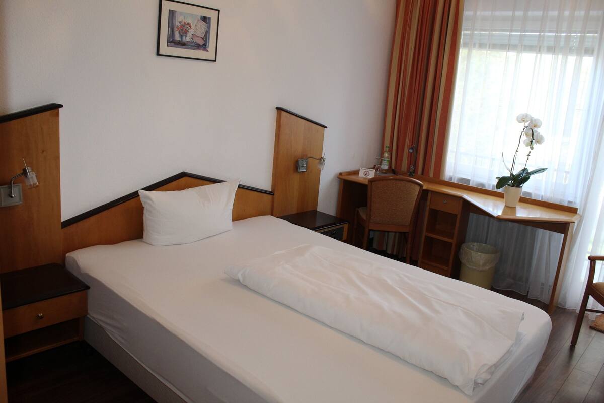 A hotel room with a bed, a desk, a chair, a trash bin, and a window with curtains.