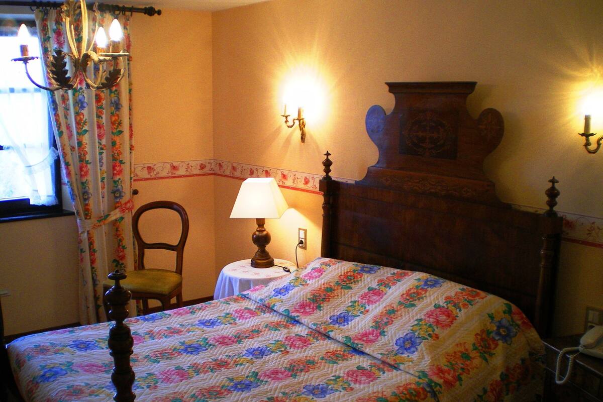 Un dormitorio con colcha estampada, cabecera de madera, una silla, una mesa con lámpara y una lámpara de pared.