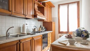 A kitchen with wooden cabinets, a stainless steel sink, and a window with lace curtains. - Verdi (Florence)