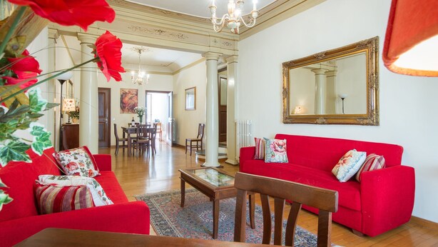 A spacious living room with a red sofa set, a glass coffee table, and a chandelier. - Verdi (Florence)