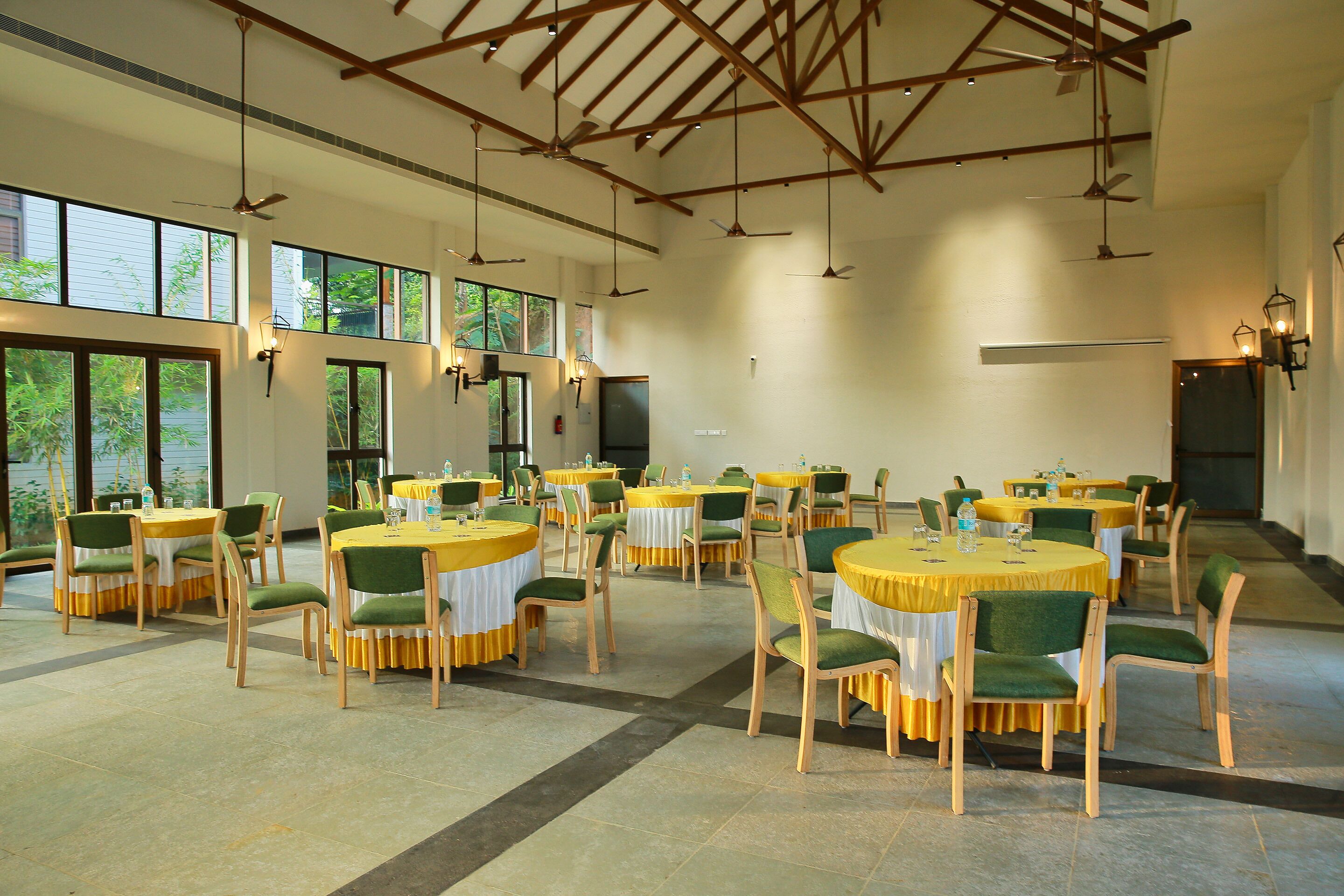 A spacious indoor dining area with multiple round tables, green upholstered chairs, and ceiling fans.