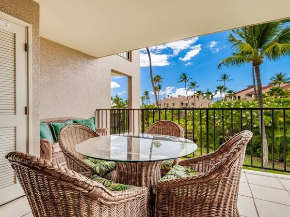 A balcony with wicker furniture and a glass table, overlooking a tropical landscape with palm trees and buildings.