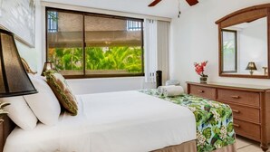 A bedroom with a large bed, a ceiling fan, a window with a view of greenery, a dresser, and a mirror.