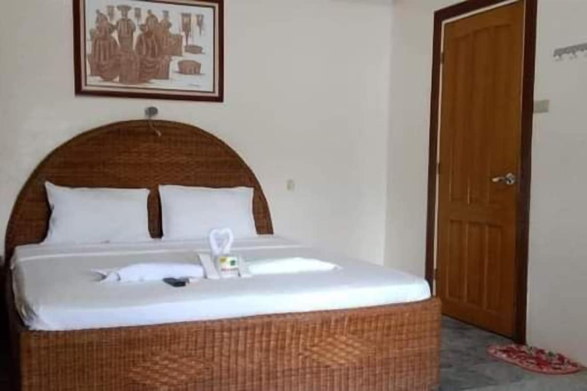 A neatly made bed with white linens and a heart-shaped pillow in a room with a wooden door and a framed picture on the wall.
