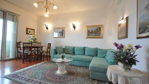 Ein Wohnzimmer mit einem türkisfarbenen Sofa, einem runden Couchtisch, einem gemusterten Teppich und einem kleinen Beistelltisch mit Blumen.