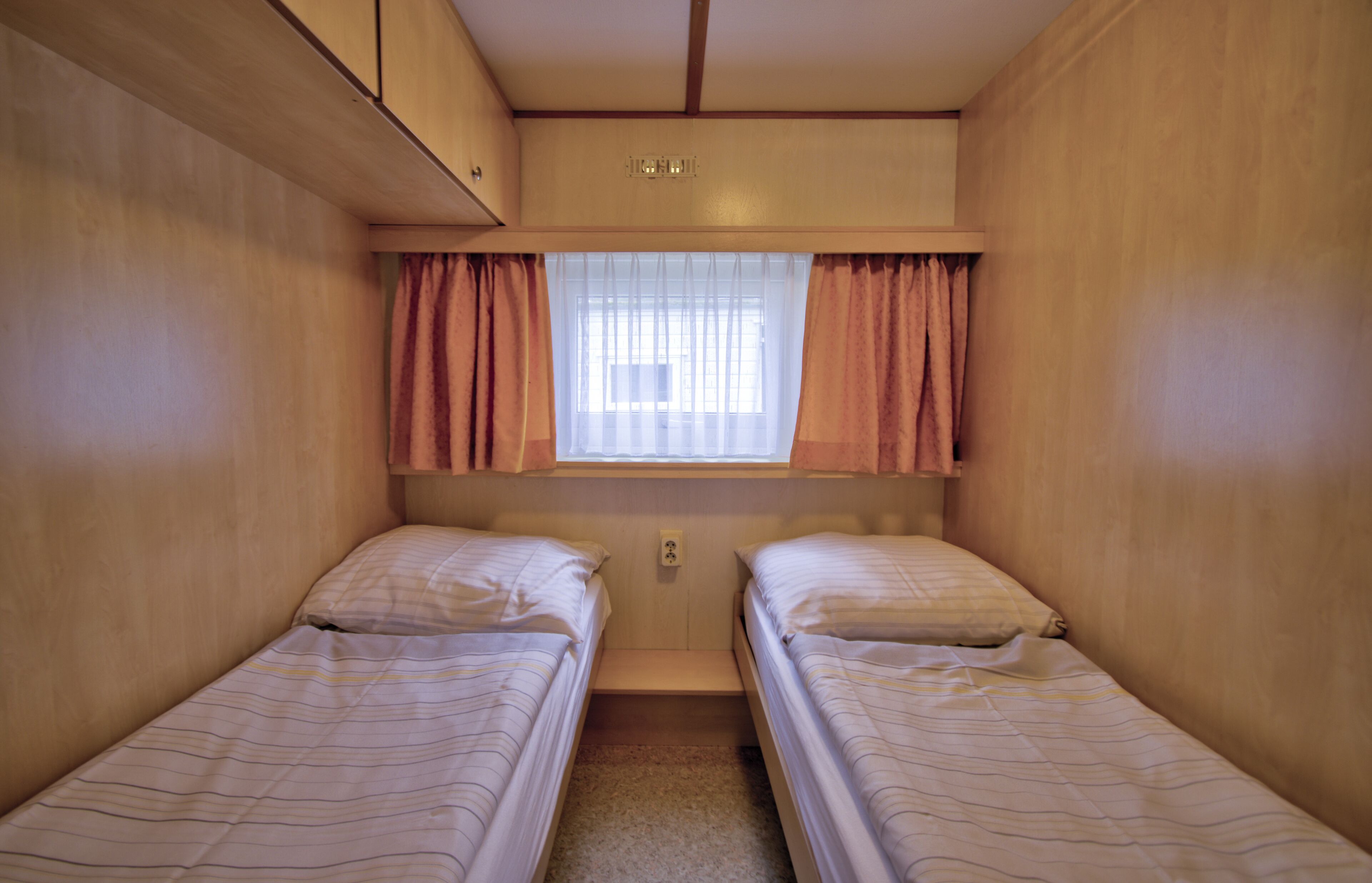 Une chambre de taille modeste, avec deux lits simples, une fenêtre avec des rideaux et une petite étagère au-dessus des lits.