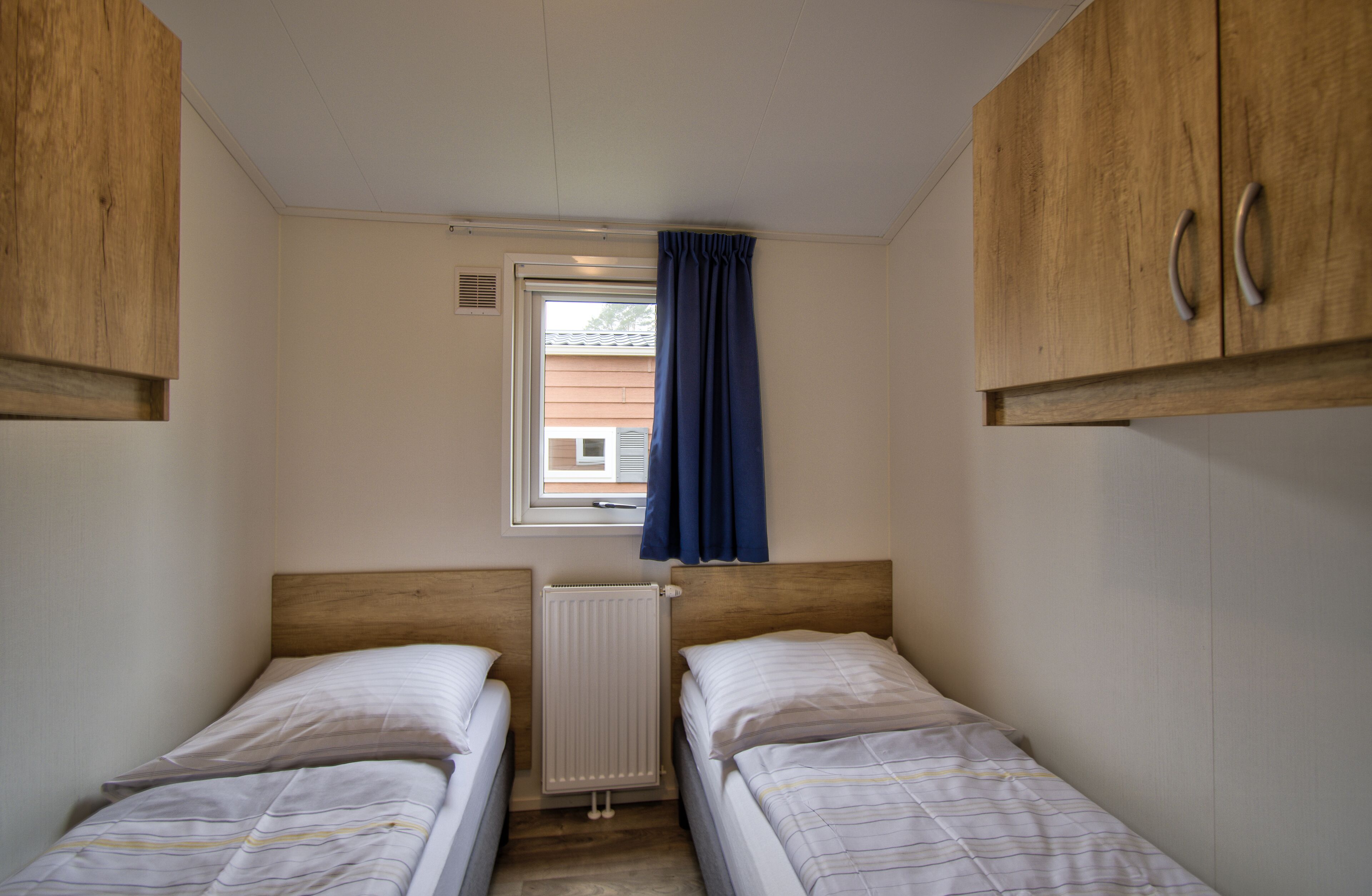 A small, compact room with two single beds, wooden cabinets, a window with blue curtains, and a radiator.
