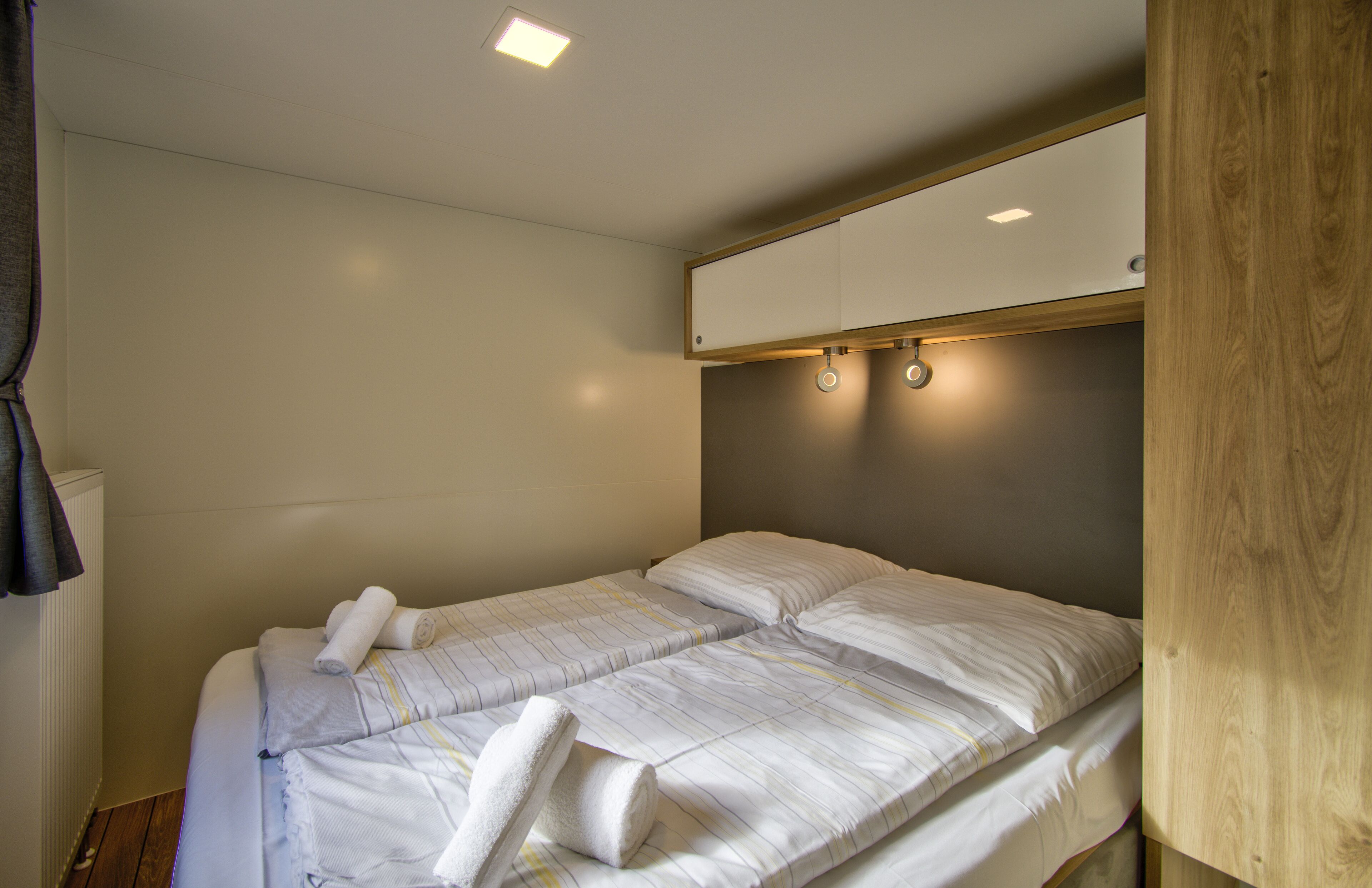 Una habitación pequeña y bien iluminada con dos camas, cabeceros de madera y una lámpara en el techo.