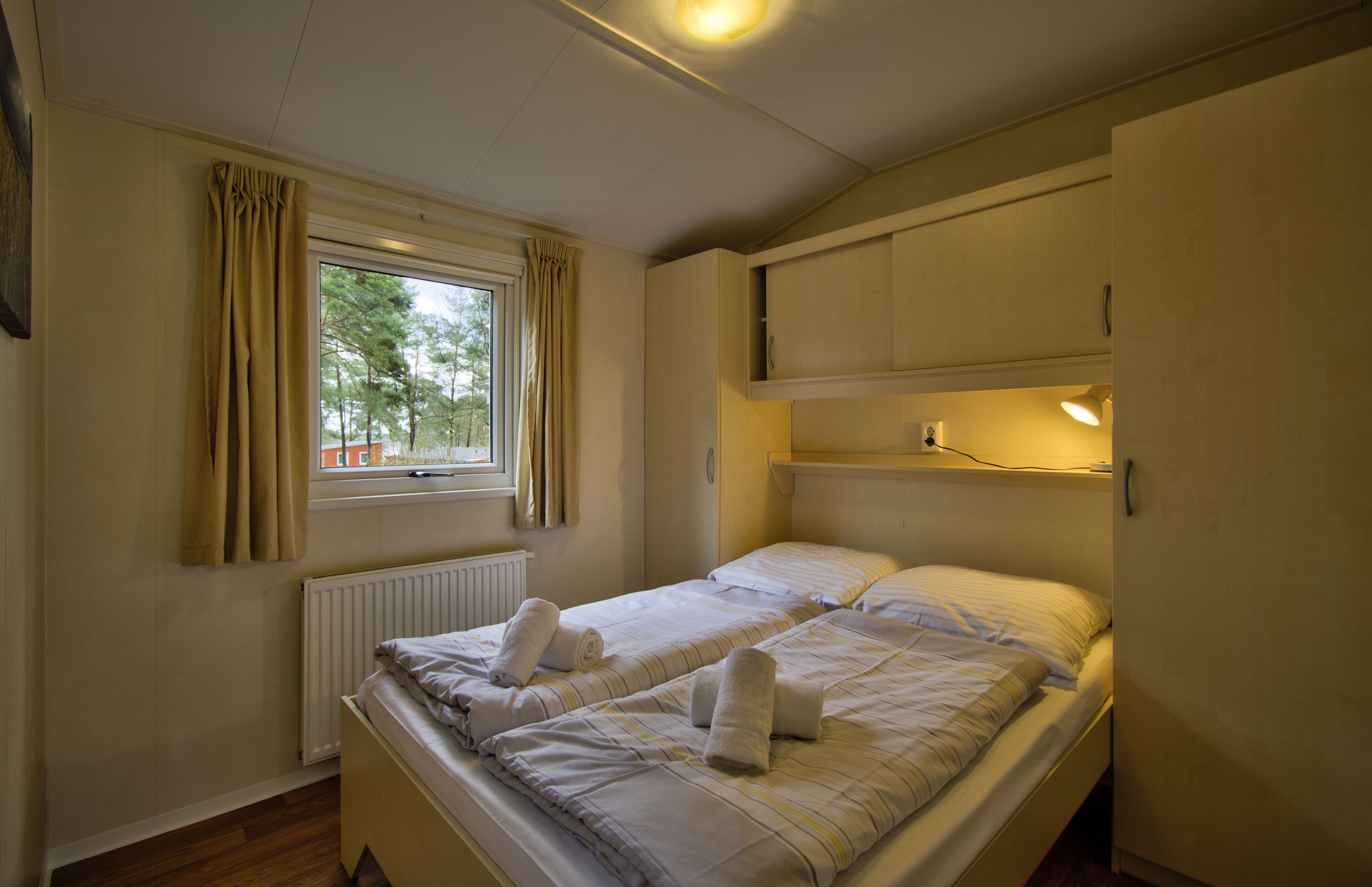 Ein Einzelbett mit zwei Kissen und einer Decke, ein Fenster mit Vorhängen, ein Heizkörper und ein Regal mit einer Lampe.