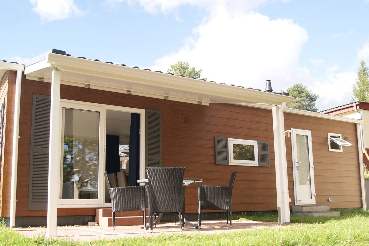 Ein Bungalow mit einer überdachten Terrasse, Schiebeglastüren und einer kleinen Sitzgelegenheit mit zwei Stühlen.