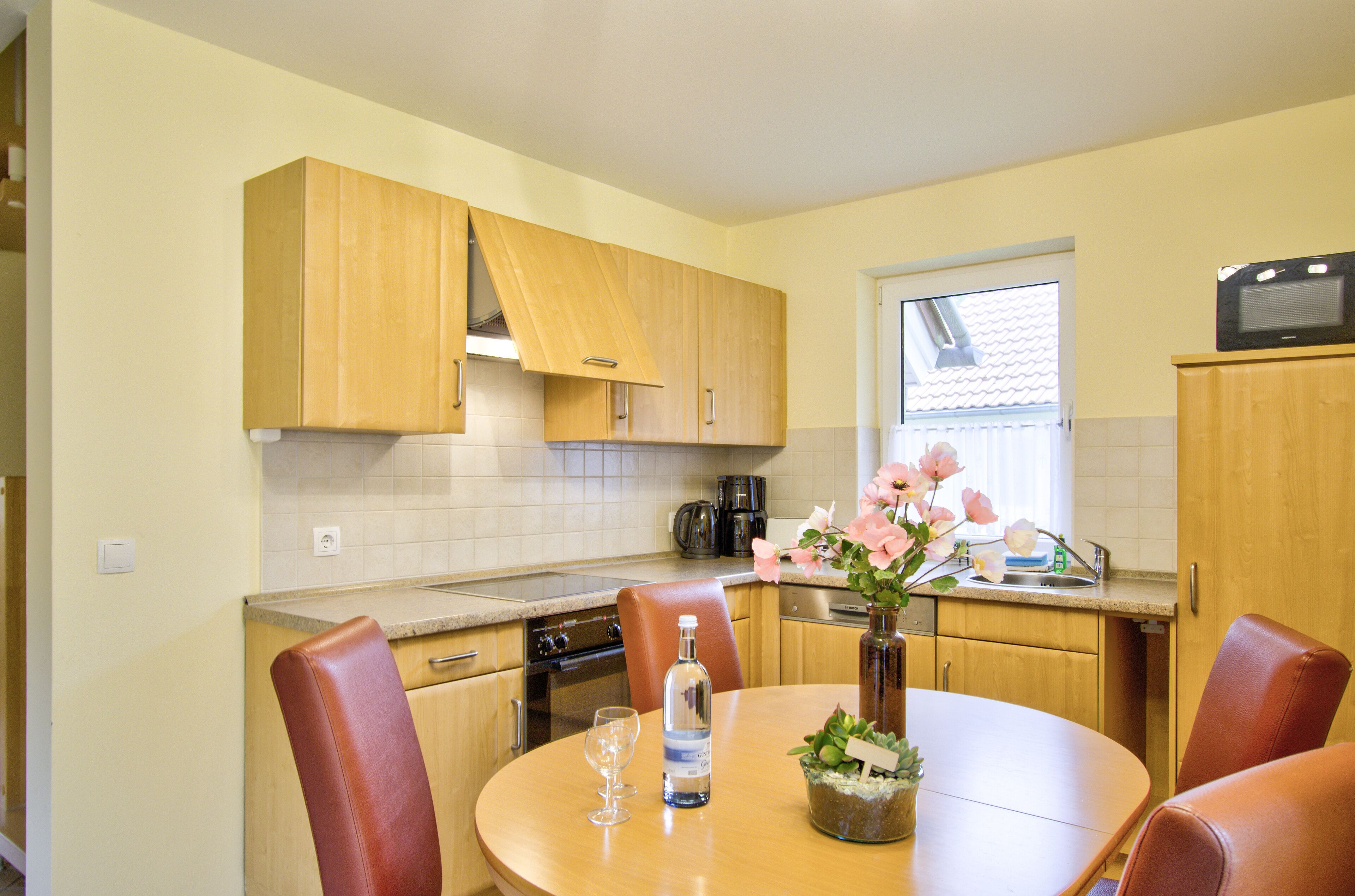 A kitchen with wooden cabinets, a dining table with chairs, a vase with flowers, a bottle, and a glass.