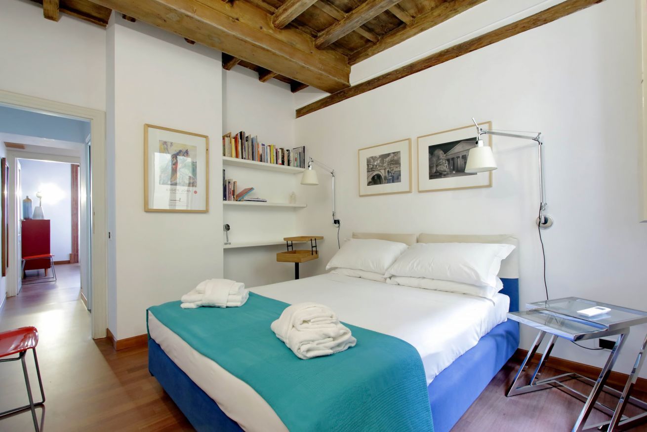 A bedroom with a bed, a desk, a chair, a bookshelf, and a door leading to another room.