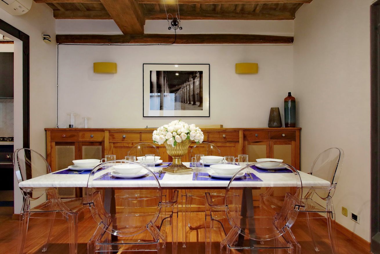 A dining area with a white table set for four, clear chairs, and a vase of white flowers.