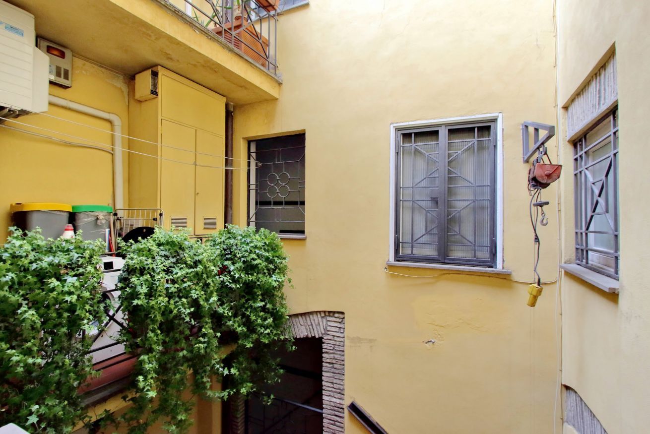 A narrow alleyway with a yellow building, a window with metal bars, and a potted plant.