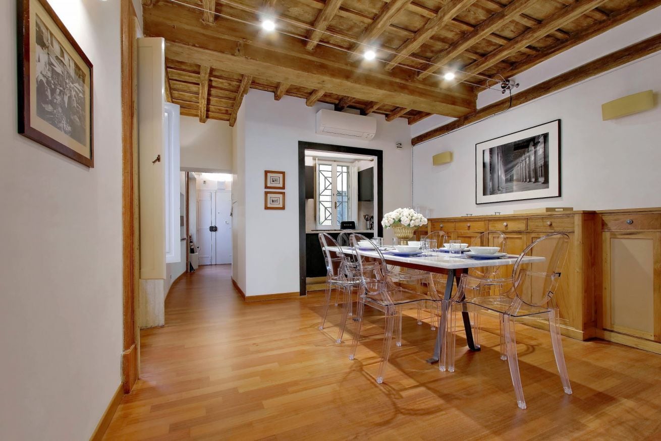 Uma área de jantar com teto de madeira, uma mesa comprida com cadeiras transparentes e quadros nas paredes.