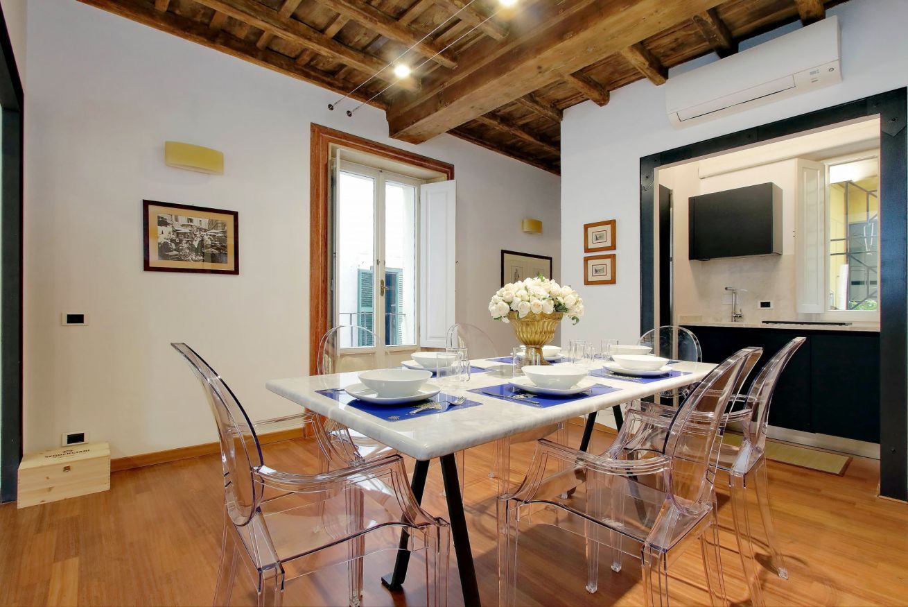 A dining area with a white table set for four, clear chairs, and a vase of white flowers.