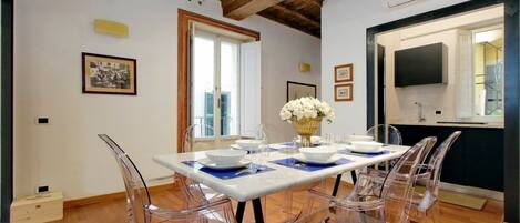 A dining area with a white table set for four, clear chairs, and a vase of white flowers.