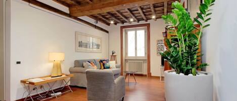 A living room with a wooden ceiling, a grey sofa, a wooden table with a lamp, and a large potted plant.