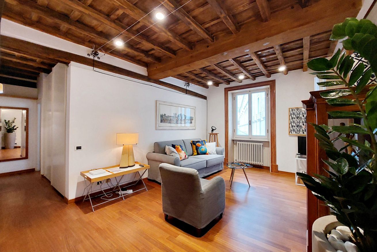 A living room with a wooden ceiling, a grey sofa, a small table with a lamp, and a large plant.