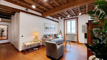 A living room with a wooden ceiling, a grey sofa, a small table with a lamp, and a large plant.