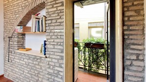 Una stanza con parete in mattoni, una mensola in legno con libri e una porta che dà su un balcone con cancello in metallo.