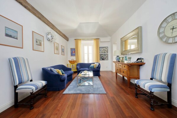 Una sala de estar con piso de madera, sofás azules, una mesa de centro de vidrio y un aparador de madera con espejo y reloj.
