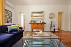 A living room with a blue sofa, a glass coffee table, a wooden dresser, a striped armchair, and a wall clock. - Superb Penthouse near Piazza Navona (Roma)