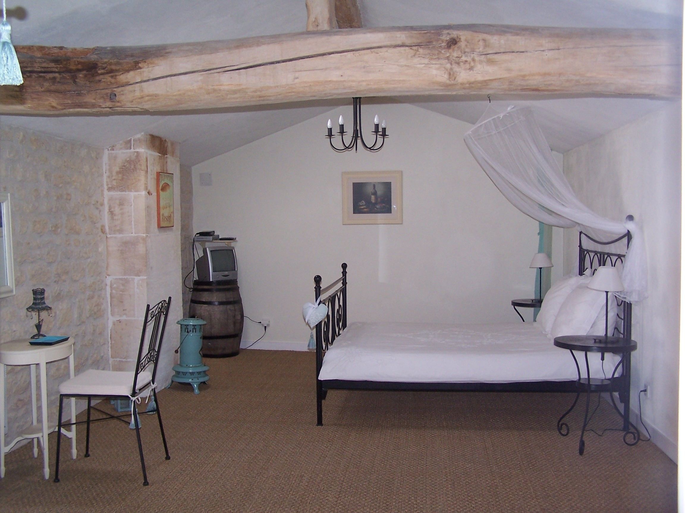 Un dormitorio con una cama, una silla, una mesita, un televisor sobre un barril y un cuadro en la pared.