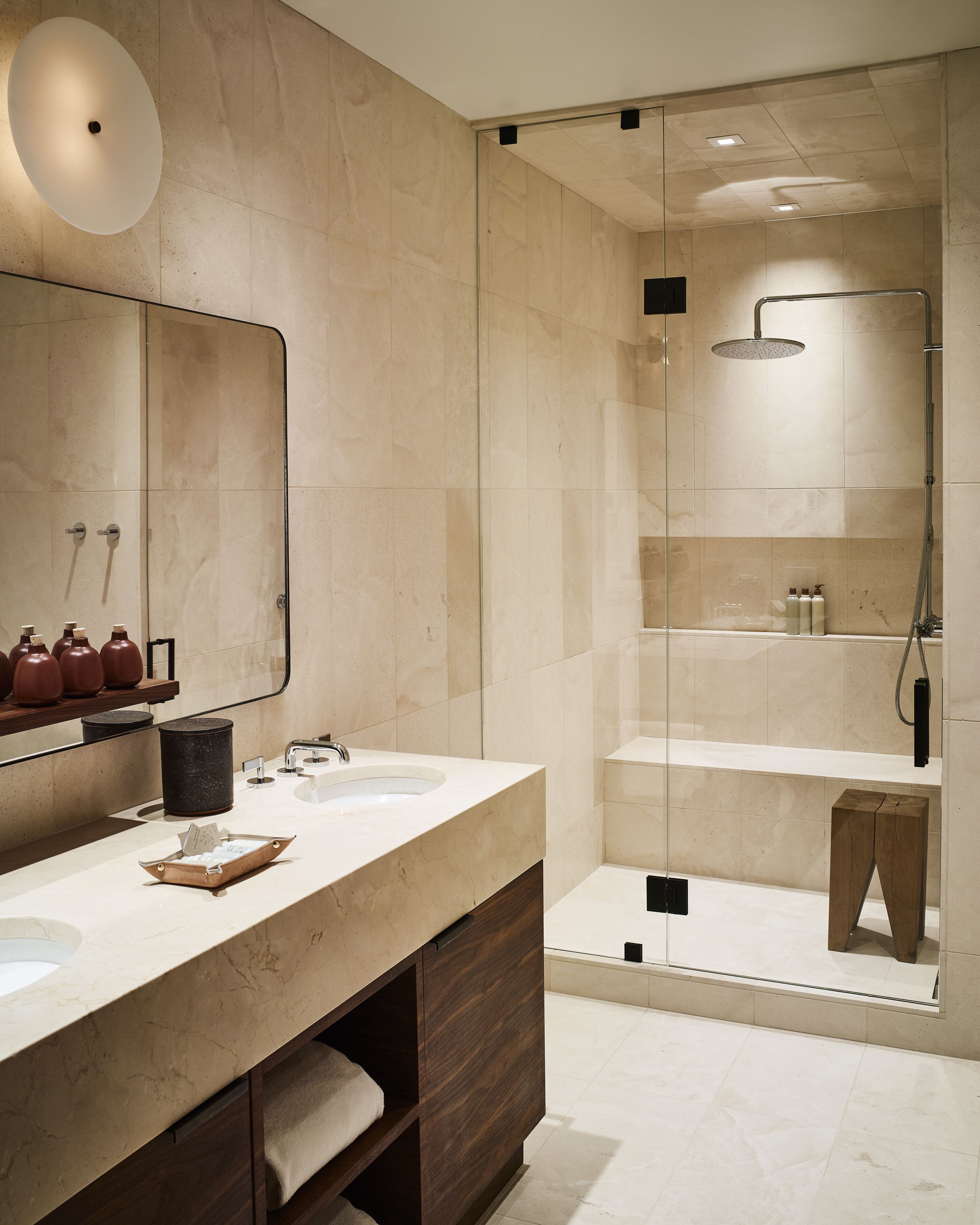 Een moderne badkamer met een grote spiegel, twee wasbakken, een douche en een houten kruk.