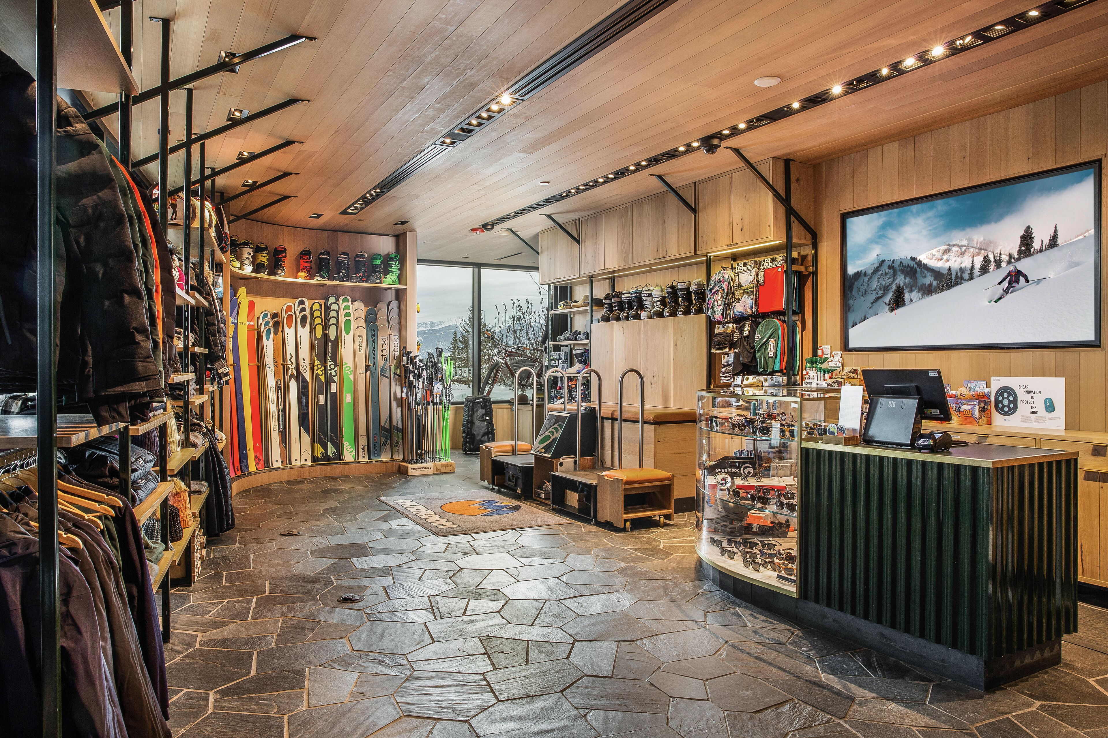 Interior de una tienda de esquí con percheros llenos de chaquetas, esquíes y equipo de esquí, un mostrador de recepción y un amplio ventanal con vistas a las montañas.