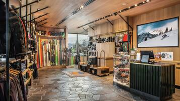 Interior de una tienda de esquí con percheros llenos de chaquetas, esquíes y equipo de esquí, un mostrador de recepción y un amplio ventanal con vistas a las montañas.