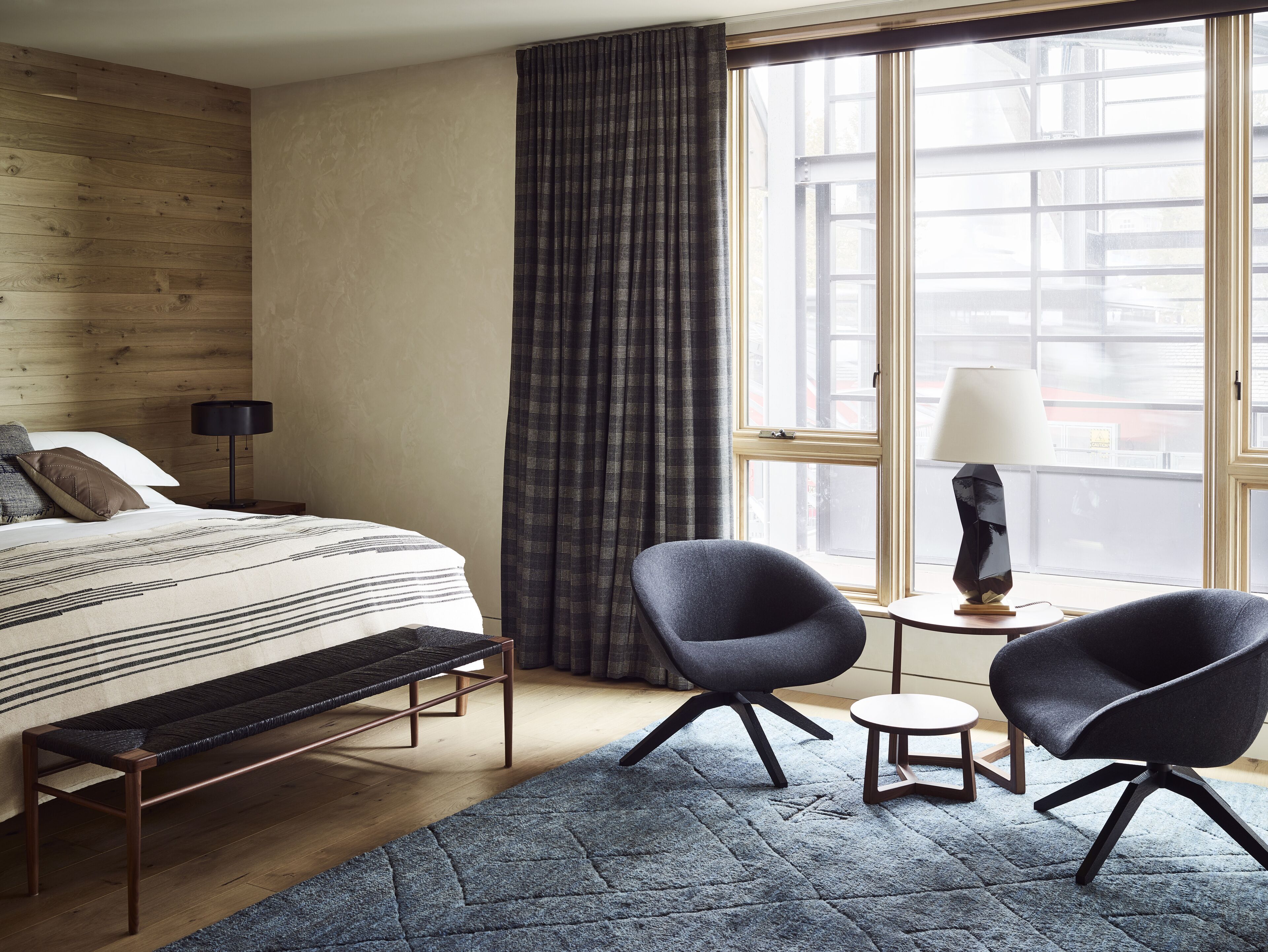 A hotel room with a bed, two armchairs, a small table, a lamp, and a large window with curtains.