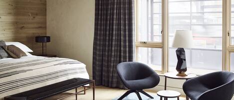 A hotel room with a bed, two armchairs, a small table, a lamp, and a large window with curtains.