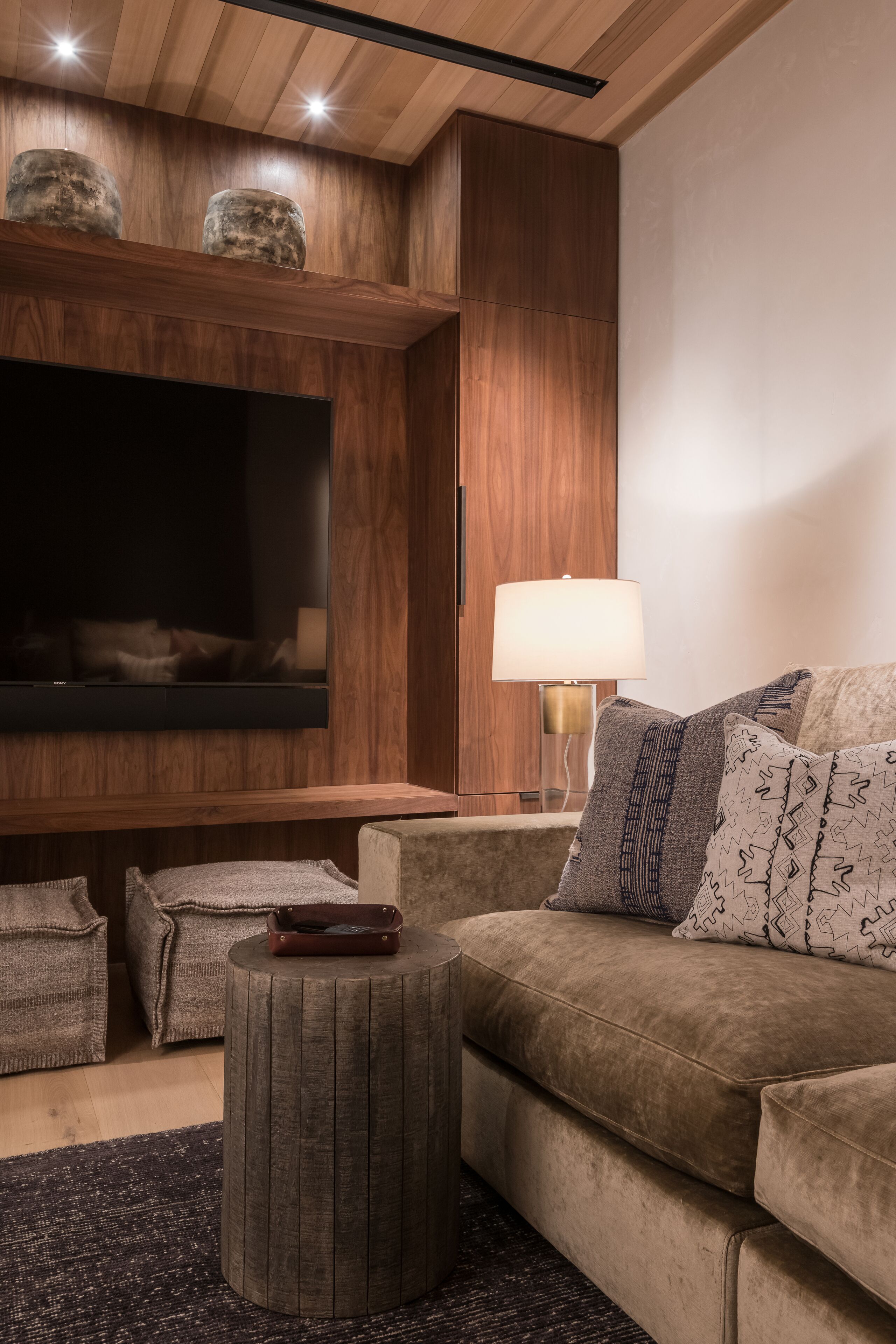 Un salon moderne doté d’un grand téléviseur à écran plat, de lambris en bois, d’un pouf rond et d’un canapé agrémenté de coussins à motifs.