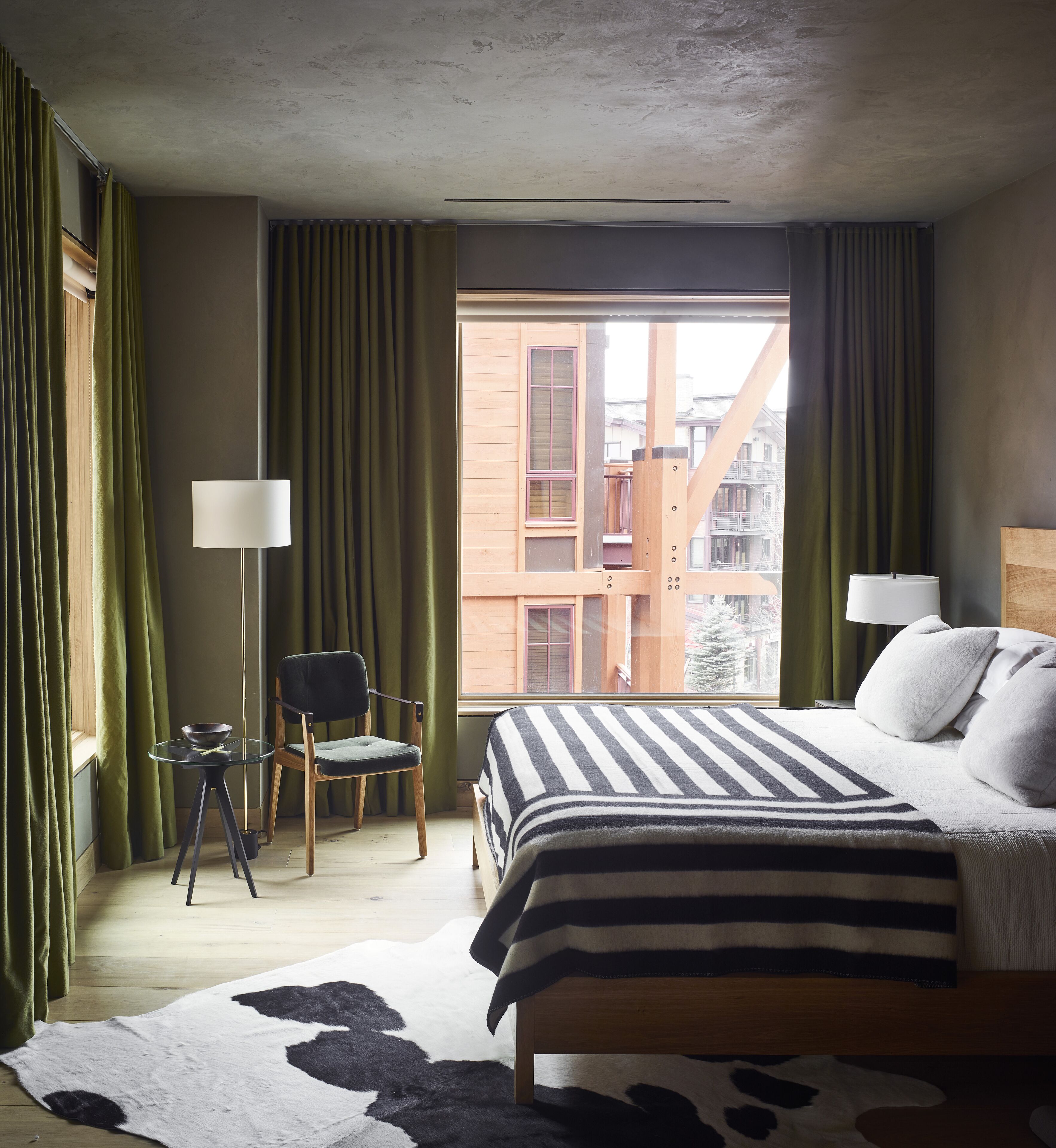 A hotel room with a bed, a chair, a table, and a view of buildings outside.