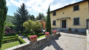A stone pathway leading to a yellow building with a door and windows, surrounded by a well-maintained garden with hedges and potted plants. - Villa La Fonte for 12 (San Godenzo)