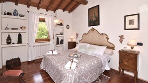 A bedroom with a large bed, wooden headboard, bedside tables, a nightstand, a window with curtains, and a view of greenery outside. - Villa La Fonte for 12 (San Godenzo)