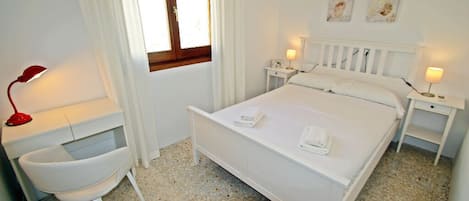 Un dormitorio con cama, mesitas de noche, una silla y una ventana con cortinas.