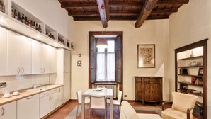 Une cuisine avec des meubles blancs, une table à manger et des chaises. On y trouve également un radiateur, un meuble en bois et une bibliothèque remplie de livres et d’objets décoratifs.