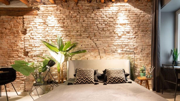 A bedroom with a brick wall, a bed with patterned pillows, a potted plant, and a modern chair. - Officine Cavour Apartments (Padova)