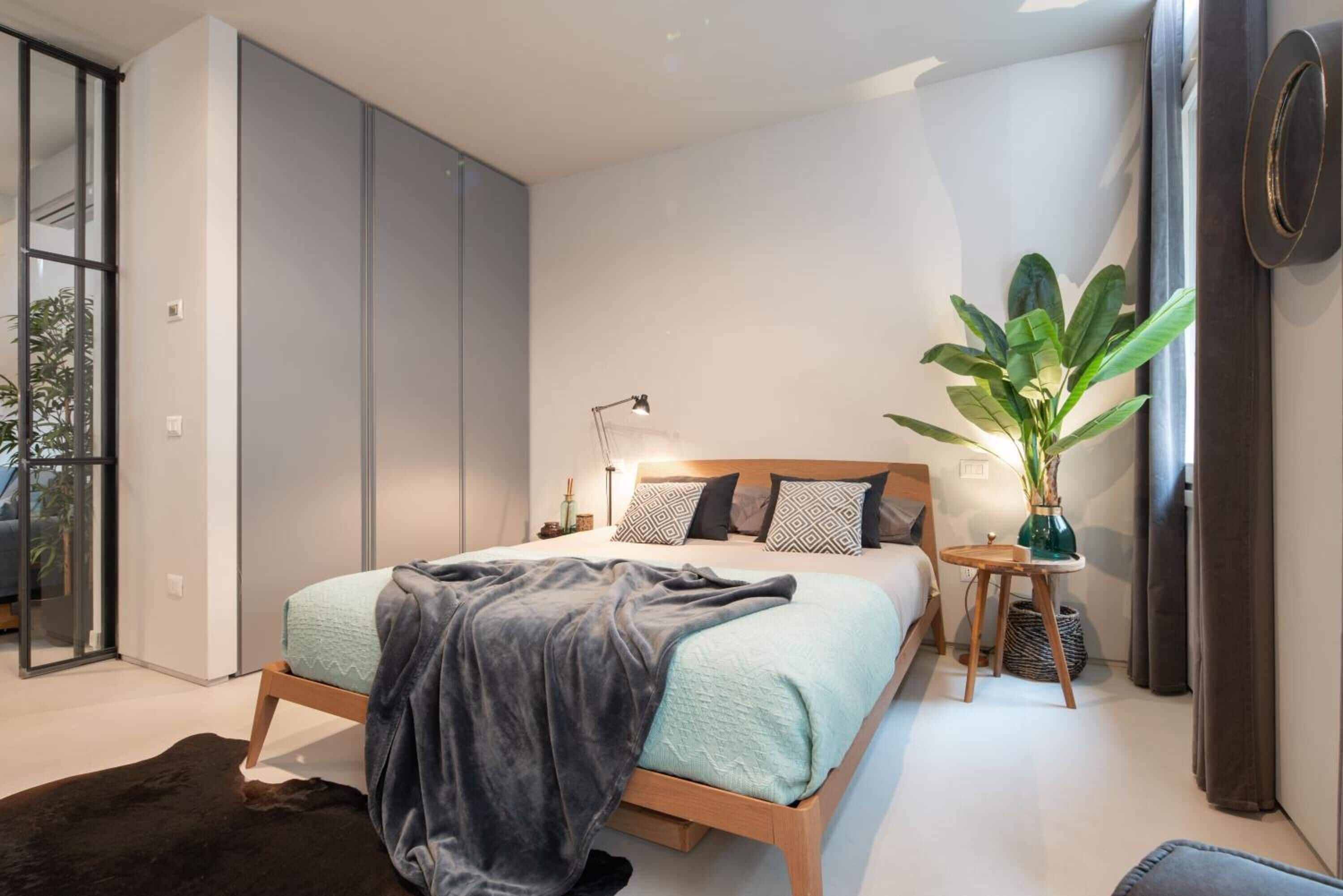 A modern bedroom with a large bed, a bedside table, a floor lamp, and a potted plant.