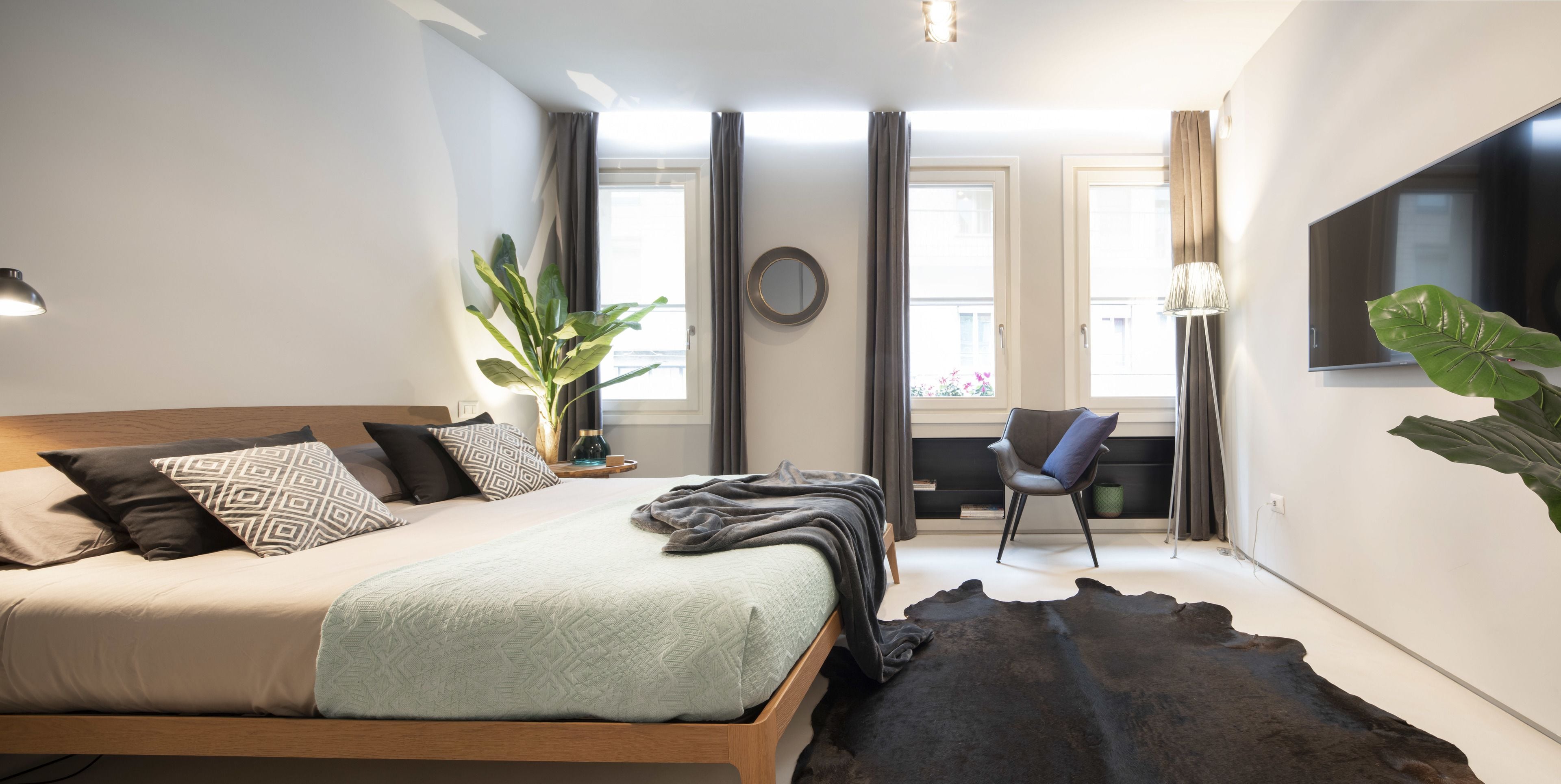 Une chambre moderne avec un grand lit, un téléviseur fixé au mur, un fauteuil et une grande plante.
