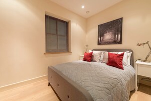 Un dormitorio con cama, mesita de noche, lámpara, ventana con persianas y un cuadro colgado en la pared.