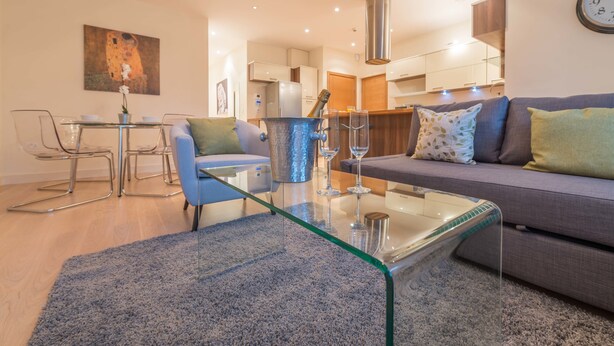 A modern living room with a glass coffee table, a sofa with patterned cushions, and a dining area with a metal chair and a vase of flowers. - Hampden Apartments - The Catherine (Windsor)